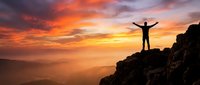Male hiker with raised arms celebrating success on mountain peak at dramatic orange sunset with layered mountain ranges and golden sky for motivation.