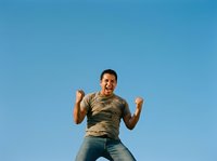 Man in casual khaki shirt and jeans jumping with excitement against clear blue sky. Male adult, South Asian ethnicity, expressing joy and enthusiasm. Suitable for lifestyle, motivation, and happiness concepts.