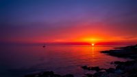 Dramatic sunset over calm ocean waters with golden sun reflecting on sea surface, silhouetted sailboat and rocky coastline, vibrant purple and orange sky for travel and nature backgrounds.