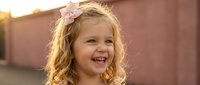 Young blonde girl with curly hair and pink flower headband smiling joyfully outdoors during golden hour sunset light creating warm portrait.