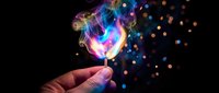 Glowing light bulb with colorful smoke and sparkling particles held by hand against dark background symbolizing creativity and innovation.
