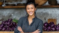 Young Asian female chef smiling confidently with arms crossed in kitchen, wearing dark apron against backdrop of purple cabbages and rustic shelving.