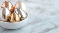 Metallic Easter eggs in rose gold, silver and gold finish nestled in white ceramic bowl on marble surface, modern minimal holiday composition.