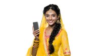 Young South Asian woman in traditional yellow and pink wedding attire with jewelry and bindi holds smartphone while smiling. Isolated on white background for design use.