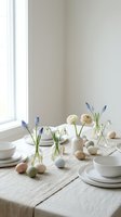 Minimalist Easter table setting with pastel eggs, white dishes and spring flowers on linen tablecloth near window for holiday dining and seasonal decor.