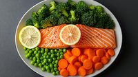 Fresh salmon fillet with lemon slices served on white plate with steamed broccoli, green peas and sliced carrots. Healthy protein-rich dinner meal.