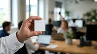 Man in white shirt holding blank white business card in modern office environment with colleagues working in background.