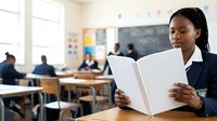 Female teacher of African descent reading an open book in a classroom with students and educational materials visible in the background during lesson preparation.