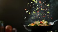 Asian stir fry noodles with fresh vegetables and herbs tossed in wok with flying ingredients against dark background, dramatic food photography.
