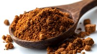 Ground aromatic cinnamon powder in wooden spoon with scattered spice crumbs on white surface, macro food photography for cooking and baking.