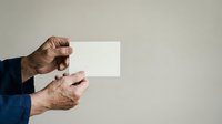 Senior man with weathered hands holding blank white card against neutral background, wearing dark blue sleeve. Suitable for business messaging, testimonials, announcements, and promotional campaigns.