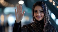 Young Middle Eastern woman wearing black lace hijab waves hello with welcoming smile against soft bokeh lights background, representing diversity and inclusion.
