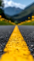 Yellow road marking line on asphalt highway stretches into distance with blurred mountain landscape and wildflowers, shot from ground level perspective with shallow depth of field.