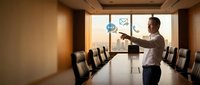 Young male professional in business casual attire presenting digital communication concept in modern conference room with large window and empty chairs for meetings.