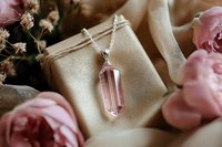 Pink crystal pendant necklace on silver chain with rose quartz point, surrounded by soft pink roses and delicate dried flowers on beige fabric background.