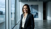 Middle-aged woman with dark hair and Mediterranean ethnicity wearing professional black blazer and white shirt standing in modern high-rise office with floor-to-ceiling windows overlooking cityscape. Corporate executive portrait for business, leadership, and professional services.