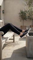 Woman's legs in black leggings and gray slip-on sneakers resting on a wooden bench in a sunny outdoor patio with potted olive trees.