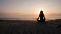 Woman meditating in lotus pose on beach at sunset with golden sky and ocean waves creating peaceful mindfulness moment for wellness and spiritual practice.