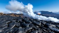 Active volcano eruption with lava flow and billowing white smoke against blue sky, viewed from rocky volcanic terrain.