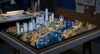 Detailed architectural scale model of modern waterfront city development with skyscrapers, marina, and urban planning layout displayed on table in office setting.