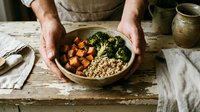 Man holding wooden bowl with roasted sweet potato cubes, kale and quinoa grain for healthy lunch meal preparation on rustic table.