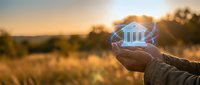 Hand holding glowing holographic bank building icon at sunset representing digital banking, fintech innovation and future financial services technology.