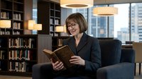 Professional middle-aged Caucasian businesswoman with glasses reading book in modern office library with city view, corporate learning concept.