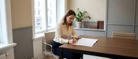 Professional businesswoman working at wooden desk in modern office space reviewing documents and paperwork for corporate meeting preparation.