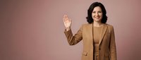 Professional businesswoman in beige suit waving hello with friendly smile on pink background for corporate presentations and marketing materials.