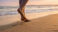Woman walking barefoot on sandy beach at sunset leaving footprints in warm golden light with ocean waves in background for wellness and travel concepts.