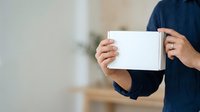 Person in dark navy shirt holding blank white envelope or card with both hands, displaying front-facing mockup space for branding, messaging, or promotional content in indoor setting.
