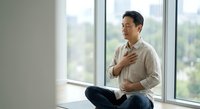 Asian man sitting cross-legged on yoga mat, hands on chest and belly, practicing mindful breathing meditation near bright window.