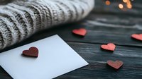 Romantic composition with white envelope, red paper hearts and cozy knitted sweater on dark wooden background. Soft bokeh lights create warm atmosphere.