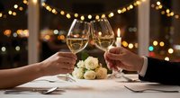 Couple toasting with white wine glasses at romantic dinner with roses and warm string lights creating intimate restaurant atmosphere for celebration.