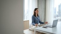 Young Asian businesswoman meditating at modern office desk with laptop and city view, practicing mindfulness and stress relief during work break.