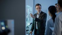Middle-aged male professional wearing business suit and glasses presenting data visualization on digital screen to diverse team in modern office environment.