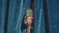 Woman hand holding champagne flute with golden bubbles through blue fabric curtains for celebration and luxury lifestyle concepts.