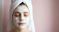 Young woman with white clay facial mask and towel wrapped around head relaxing with closed eyes during spa skincare treatment routine.