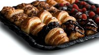 Fresh baked mini croissants and pastries arranged on black catering tray with chocolate drizzle, berries and nuts, close-up view with selective focus.