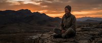 Young man meditating in lotus position on rocky desert landscape during golden hour sunset with dramatic mountain silhouettes and warm orange sky creating peaceful atmosphere.