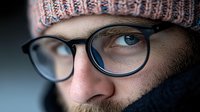 Close caucasian man wearing glasses and knit winter hat, with intense blue eyes looking directly at camera. Shallow depth of field.