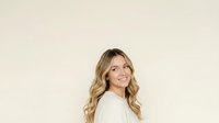Young woman with long blonde wavy hair and fair complexion, wearing a light cream-colored shirt, smiling while looking upward against a neutral beige background.