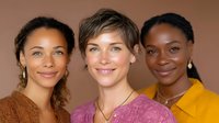 Diverse group of three smiling women with different hairstyles and ethnicities wearing casual clothing against brown background, representing friendship and inclusivity.