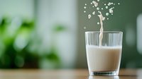 Splash of milk creating crown-shaped droplets in tall glass against green blurred background, dynamic liquid motion captured in high-speed photography.