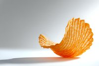 Single wavy potato chip casting soft shadow on white surface, crispy ridged texture and warm orange color create minimalist food composition.