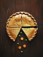 Homemade chicken pot pie with flaky golden crust cut into portions, decorated with fresh green peas and sliced carrots on dark wooden background, overhead view.