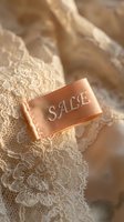 Peach satin sale tag with embroidered text resting on delicate cream lace fabric, evoking luxury fashion retail and seasonal discount promotions.
