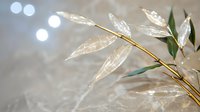 Delicate crystal leaves on golden branch with natural green foliage against soft bokeh background create ethereal winter holiday atmosphere.