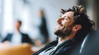 Joyful caucasian businessman laughing during meeting, sitting in office chair with genuine emotional expression. Positive workplace atmosphere and success concept.
