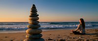 Zen stone tower on beach at sunset with woman meditating in background. Peaceful coastal scene for wellness, mindfulness and spiritual balance concepts.